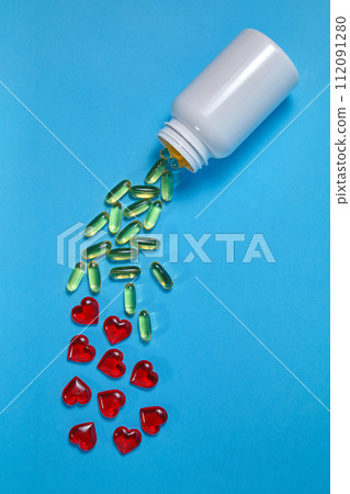 Yellow omega 3 gelatin tablets and red hearts spill out of a white jar on a blue background 112091280