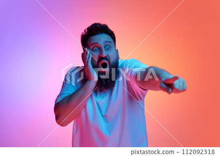 Shocked young man with widely open mouth put hand on face and points into distance in neon light against vibrant gradient studio background. Shocked young man with widely open mouth put hand on face and points into distance in neon light against vibrant gradient studio background. 112092318