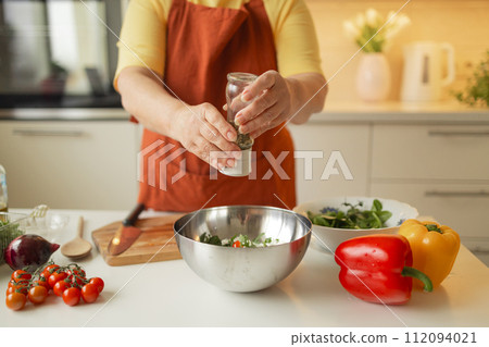 Close-up of a 60s woman preparing healthy vegetable salad in kitchen, seasoning meal, adding some herbs and salt to bowl, enjoying cooking vegetarian food at home, free space for advertising text 112094021