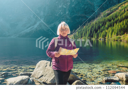 Shot of a senior 60s lost woman with smart phone holding a map while taking in the view from the top of a mountain. Female tourist in mountain read the map. Poland, Tatry  112094544