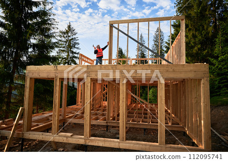 Carpenter crafting a wooden, two-story framework house. Bearded man nailing with hammer, dressed in protective hardhat and construction vest. Concept of modern, ecologically-responsible construction. 112095741