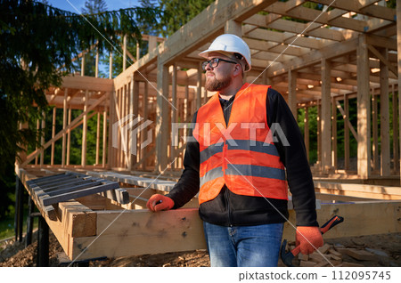 Carpenter constructing wooden frame two-story house near the forest. Bearded man holding hammer, dressed in protective helmet and orange safety vest. Concept of ecological modern construction. 112095745