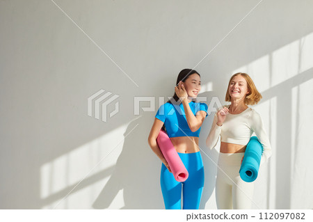 Two young women stand in yoga class, smiling and talking. Concept of physical and mental health care, asian and caucasian ethnicity Two young women stand in yoga class, smiling and talking. Concept of physical and mental health care, asian and caucasian ethnicity 112097082
