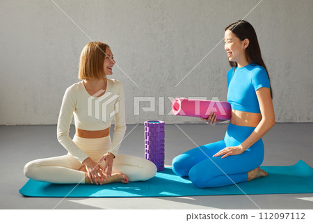 Two young women sitting in yoga class, smiling and talking. Concept of physical and mental health care, asian and caucasian ethnicity 112097112