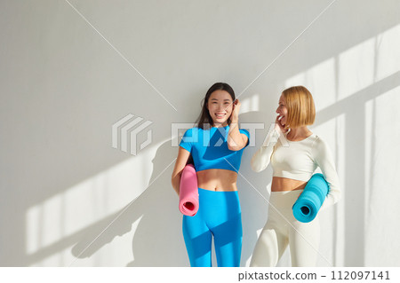 Two young women stand in yoga class, smiling and talking. Concept of physical and mental health care, asian and caucasian ethnicity 112097141