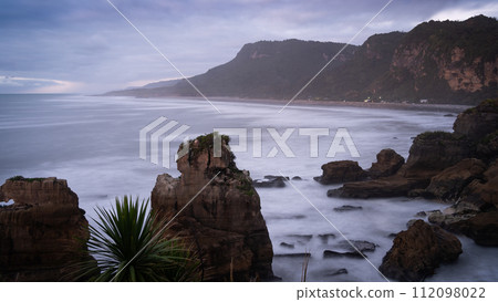 Rough ocean coast with waves and some houses and sharp cliffs during sunset, New Zealand 112098022