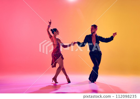 Male and female dancers performing a Latin dance in stylish clothes against gradient pink yellow background in neon light Male and female dancers performing a Latin dance in stylish clothes against gradient pink yellow background in neon light 112098235