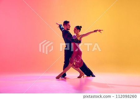 Passionate dancers. Artistic, talented young man and woman in motion in beautiful stage costumes dancing ballroom against gradient pink yellow background in neon light Passionate dancers. Artistic, talented young man and woman in motion in beautiful stage costumes dancing ballroom against gradient pink yellow background in neon light 112098260
