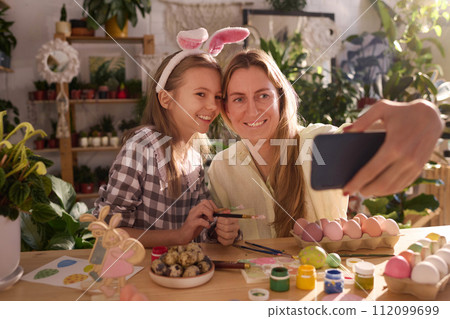Medium portrait of creative mother and daughter taking selfie on smartphone while coloring eggs for Easter 112099699