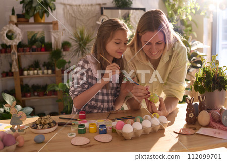 Medium shot of happy Caucasian mother and daughter decorating Easter eggs in workshop with biophilic interior 112099701