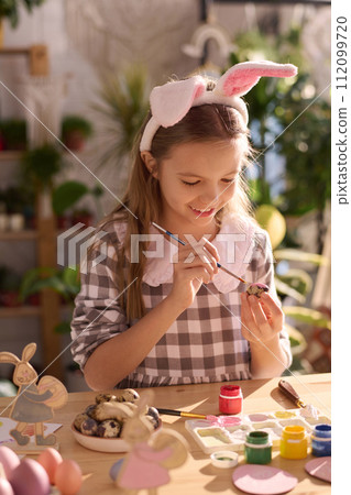 Vertical medium shot of cheerful girl wearing pretty dress with bunny ears sitting at table coloring eggs for Easter Vertical medium shot of cheerful girl wearing pretty dress with bunny ears sitting at table coloring eggs for Easter 112099720