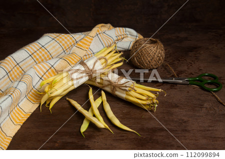 Two bunch and several pods of flat runner bean pods heap with yellow kitchen towel, scissors and ball of thread on wooden background. . 112099894