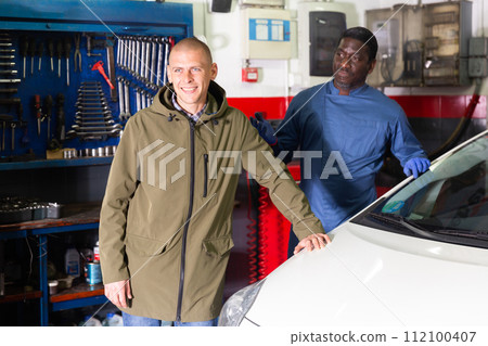 Portrait of satisfied male client with mechanician standing near her car after repairing in workshop 112100407