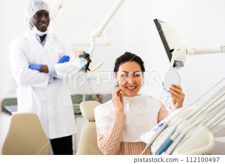 Satisfied asian woman looking in the mirror at the cured teeth at dentist Satisfied asian woman looking in the mirror at the cured teeth at dentist 112100497