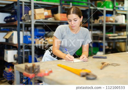 Young woman storehouse worker standing at workbench and making notes Young woman storehouse worker standing at workbench and making notes 112100533