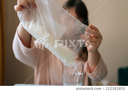 Front view of unrecognizable female artisan pouring dry mixture or soy wax from transparent bag for creating candle building mixture. Process of making handmade natural scented candle at workshop. Front view of unrecognizable female artisan pouring dry mixture or soy wax from transparent bag for creating candle building mixture. Process of making handmade natural scented candle at workshop. 112101853