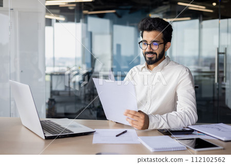 Smiling young Indian man in glasses and shirt sitting at office desk, working on laptop and documents. 112102562