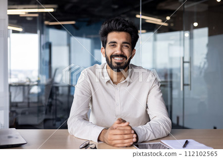 Portrait of a young Indian man in a shirt sitting smiling at a desk in the office and looking confidently at the camera. Portrait of a young Indian man in a shirt sitting smiling at a desk in the office and looking confidently at the camera. 112102565