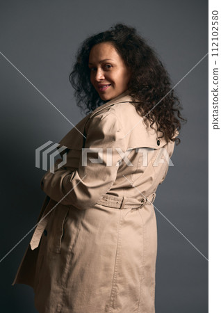 Beautiful curly haired gravid woman smiles looking at camera through her shoulder, isolated over fashion gray background Beautiful curly haired gravid woman smiles looking at camera through her shoulder, isolated over fashion gray background 112102580