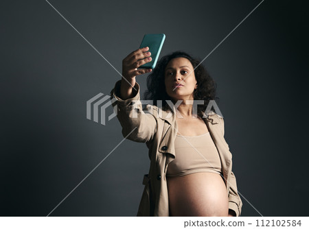 Beautiful mid adult pregnant woman with naked belly, in stylish beige trench, using mobile phone, over gray background Beautiful mid adult pregnant woman with naked belly, in stylish beige trench, using mobile phone, over gray background 112102584