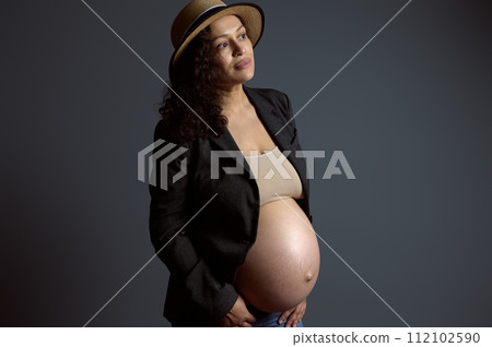 Attractive pensive young pregnant woman holding her belly, dreamily looking aside a copy space on fashion gray backdrop Attractive pensive young pregnant woman holding her belly, dreamily looking aside a copy space on fashion gray backdrop 112102590