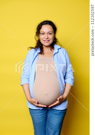 Authentic emotional portrait of a mid adult pregnant woman holding her big belly, smiling at camera. Yellow background 112102597