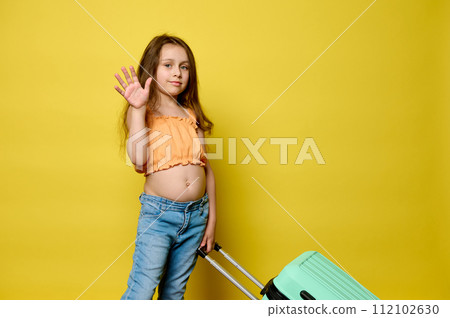 Little traveler girl carrying blue suitcase, waving hello at camera, isolated yellow background. Travel. Summer holidays Little traveler girl carrying blue suitcase, waving hello at camera, isolated yellow background. Travel. Summer holidays 112102630