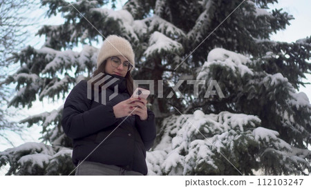 A young woman stands with a phone against a background of spruce in winter. A girl in glasses with a smartphone chats against the backdrop of snowy spruce branches. A young woman stands with a phone against a background of spruce in winter. A girl in glasses with a smartphone chats against the backdrop of snowy spruce branches. 112103247