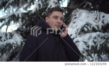A young man speaks on the phone while standing against a background of spruce in winter. A man with a phone against a background of snowy spruce branches. A young man speaks on the phone while standing against a background of spruce in winter. A man with a phone against a background of snowy spruce branches. 112103249