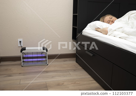 Electric Mosquito or Insect Zapper With Blue Purple Lights Turned on near Sleeping Person in Bed in Bedroom. Bug Killer Lamp on Wooden Floor in Room. Fly Trap Indoor. Horizontal Plane Electric Mosquito or Insect Zapper With Blue Purple Lights Turned on near Sleeping Person in Bed in Bedroom. Bug Killer Lamp on Wooden Floor in Room. Fly Trap Indoor. Horizontal Plane 112103377