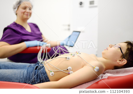 Child under ECG test with doctor. Boy patient lying in medical bed with vacuum sensors. Pediatrician looking result on tablet while heart electrocardiogram procedure at medical clinic. Child under ECG test with doctor. Boy patient lying in medical bed with vacuum sensors. Pediatrician looking result on tablet while heart electrocardiogram procedure at medical clinic. 112104393