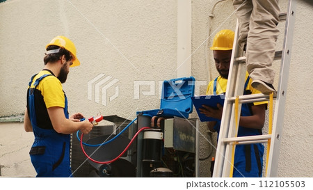 Specialists measuring freon levels in HVAC system with pressure indicators while african american coworker steps down from folding ladder after finishing checking rooftop condenser Specialists measuring freon levels in HVAC system with pressure indicators while african american coworker steps down from folding ladder after finishing checking rooftop condenser 112105503