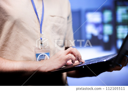 Worker in front of blurry background supercomputers delivering web content to online customers. Repairman in server room using laptop to make sure critical systems are working errorless, close up Worker in front of blurry background supercomputers delivering web content to online customers. Repairman in server room using laptop to make sure critical systems are working errorless, close up 112105511