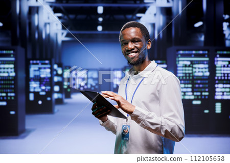 Portrait of african american administrator in front of server clusters in high performance computing data center. Technician monitoring energy consumption across systems, ensuring optimal operations Portrait of african american administrator in front of server clusters in high performance computing data center. Technician monitoring energy consumption across systems, ensuring optimal operations 112105658