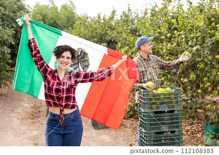 Excited Latin woman holding flag of Mexico while workers picking lemons on farm 112108383