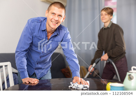 Father cleans the table with a rag while son vacuums room 112108504