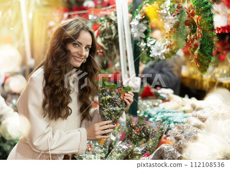 Smiling woman choosing festive home decorations on street Christmas fair Smiling woman choosing festive home decorations on street Christmas fair 112108536