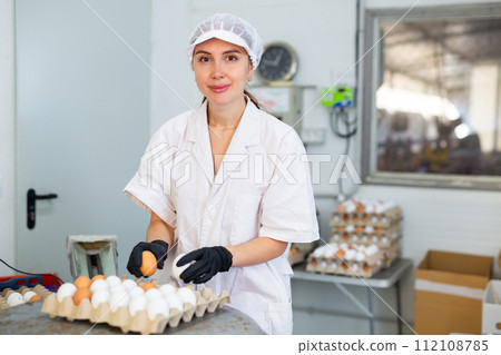 Focused young woman farmer worker in white coat sorting and labeling chicken eggs, putting eggs to special trays on farm Focused young woman farmer worker in white coat sorting and labeling chicken eggs, putting eggs to special trays on farm 112108785