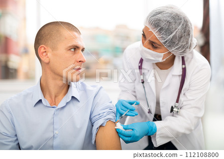Doctor in a protective mask injects a vaccine to a man with syringe Doctor in a protective mask injects a vaccine to a man with syringe 112108800