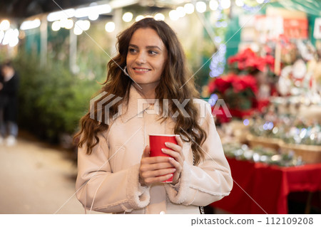 Smiling woman walking through Christmas street market with warm beverage in paper cup Smiling woman walking through Christmas street market with warm beverage in paper cup 112109208