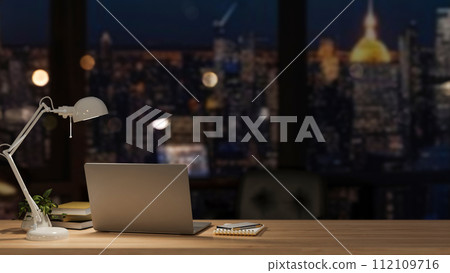 A modern office at night with a city view, featuring a laptop and accessories on a wooden desk. 112109716