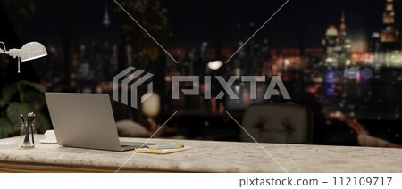 A modern office at night with a city view, featuring a laptop and accessories on a desk. 112109717