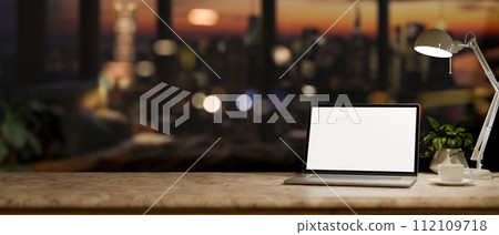 An office desk with a white-screen laptop computer in a modern dark office at night. 112109718