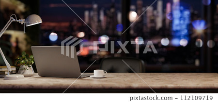 A modern office at night with a city view, featuring a laptop and accessories on a desk. 112109719