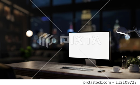 A computer desk in a modern dark office at night with a city view through the large glass window. 112109722