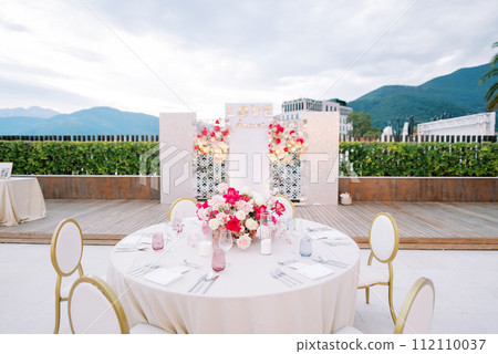 Round festive table with chairs stands opposite a stand with bouquets of flowers. Caption: Love forever Round festive table with chairs stands opposite a stand with bouquets of flowers. Caption: Love forever 112110037