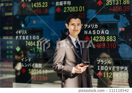 A man in his 30s smiling and looking at the camera in front of an electronic bulletin board displaying stock prices from around the world (strategy image) 112110542