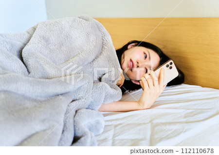 A woman in her 30s who overslept due to lack of sleep or poor physical condition after waking up in the morning and looking at her smartphone while lying on the bed in her bedroom. A woman in her 30s who overslept due to lack of sleep or poor physical condition after waking up in the morning and looking at her smartphone while lying on the bed in her bedroom. 112110678