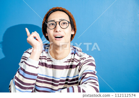 一位戴著眼鏡、戴著針織帽的年輕人坐在藍色背景的椅子上思考 112112574