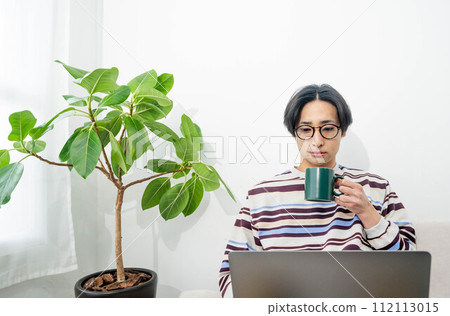 一個男人放鬆地坐在沙發上喝咖啡並操作電腦 一個男人放鬆地坐在沙發上喝咖啡並操作電腦 112113015